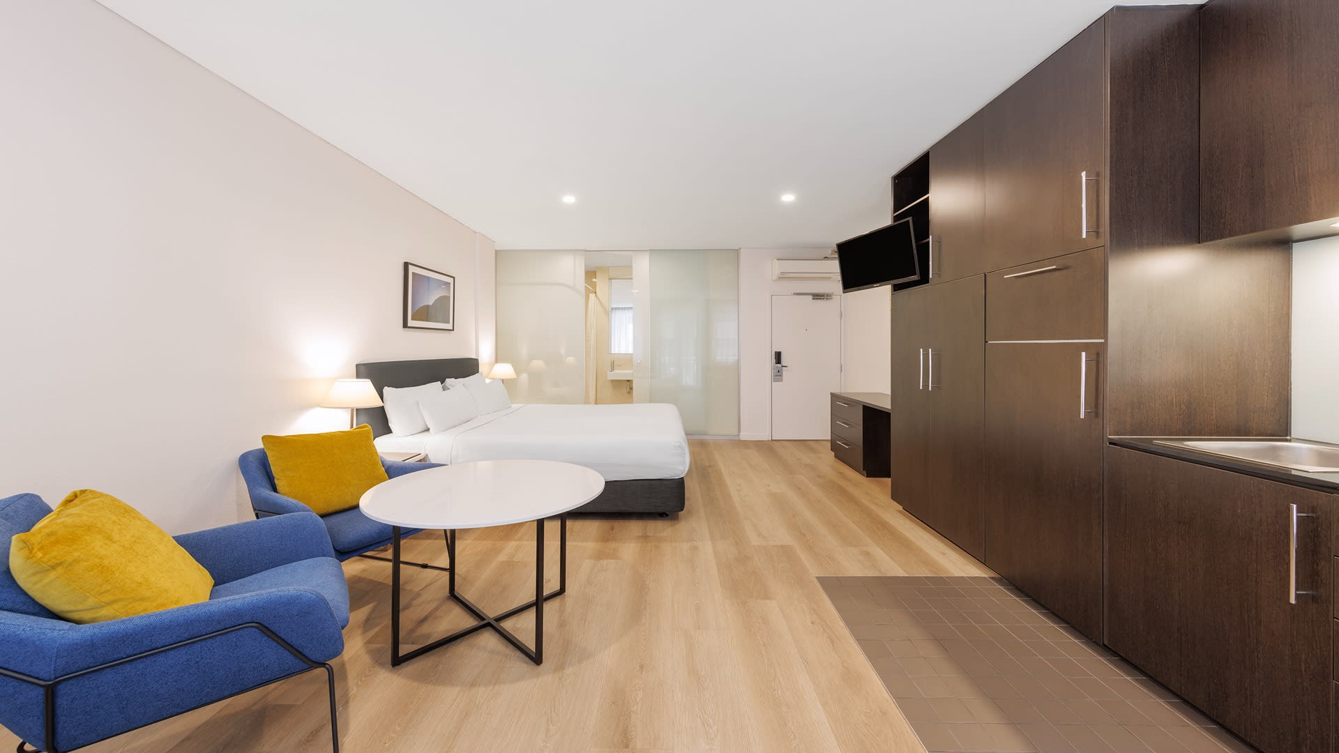 Modern studio accommodation near AO Melbourne featuring a queen bed, two blue chairs, a dining table and dark timber kitchenette.