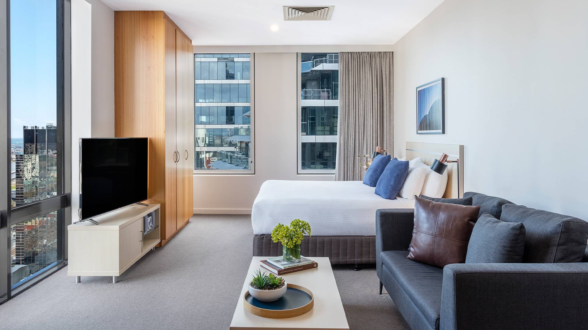 Modern studio accommodation with a king bed, grey sofa, TV, coffee table and floor-to-ceiling city views.