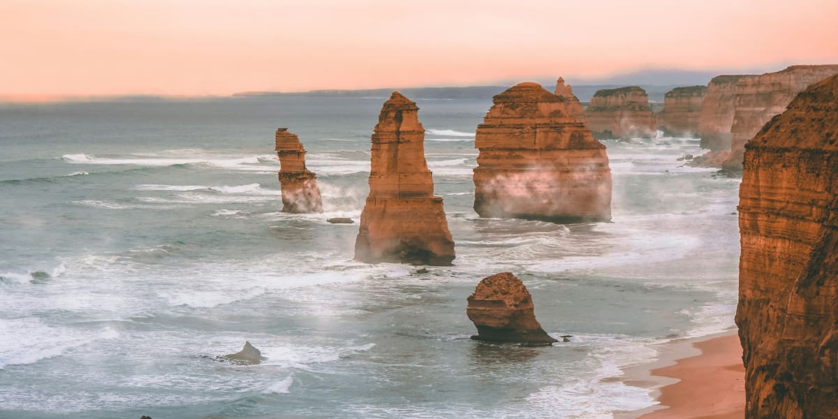 Limestone sea stacks rise from rolling ocean waves beside rugged cliffs and a sandy beach under a soft pink sky.