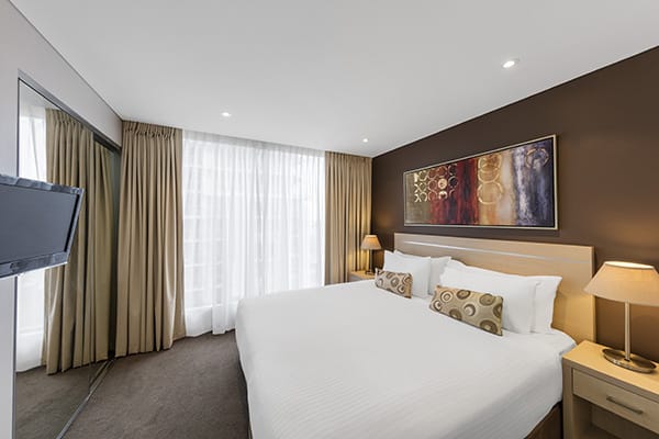 Hotels Adelaide CBD with sunlight pouring through curtains into 2 bedroom hotel apartment with queen size bed, satellite TV and pictures on walls