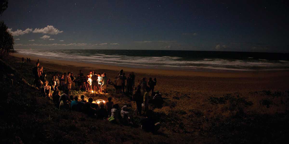Group of friends wrapped around a bonfire in the middle of the night watching the hatchlings of baby turtles in Mon Repos