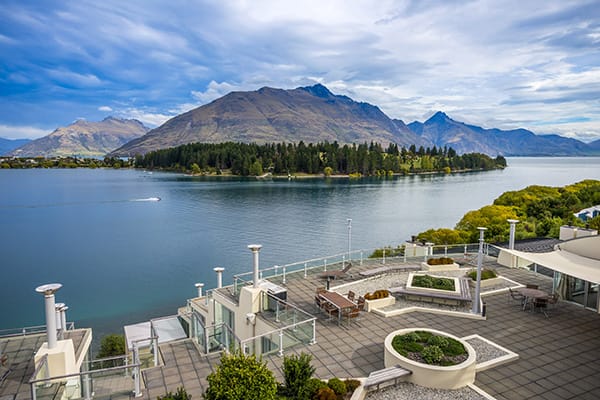 hotel guests in large courtyard of Oaks Club Resort next to Lake Wakatipu in Queenstown with The Remarkables mountain range in background during midday