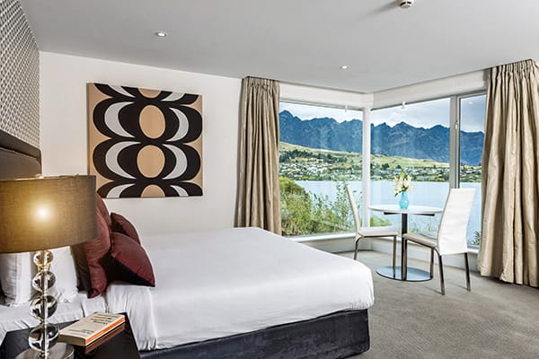 comfortable double bed with fluffy pillows and clean sheets facing large windows with views of The Remarkables mountain range and Lake Wakatipu in Queenstown during summer