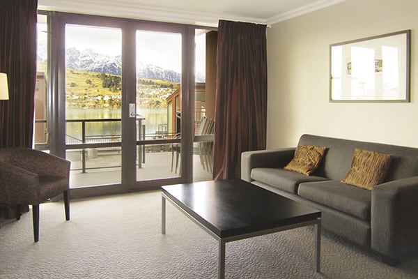 large living room with table and couches next to sliding glass doors leading to sunshine on private balcony outside 2 bedroom holiday apartment at Oaks Shores hotel in Queenstown, New Zealand